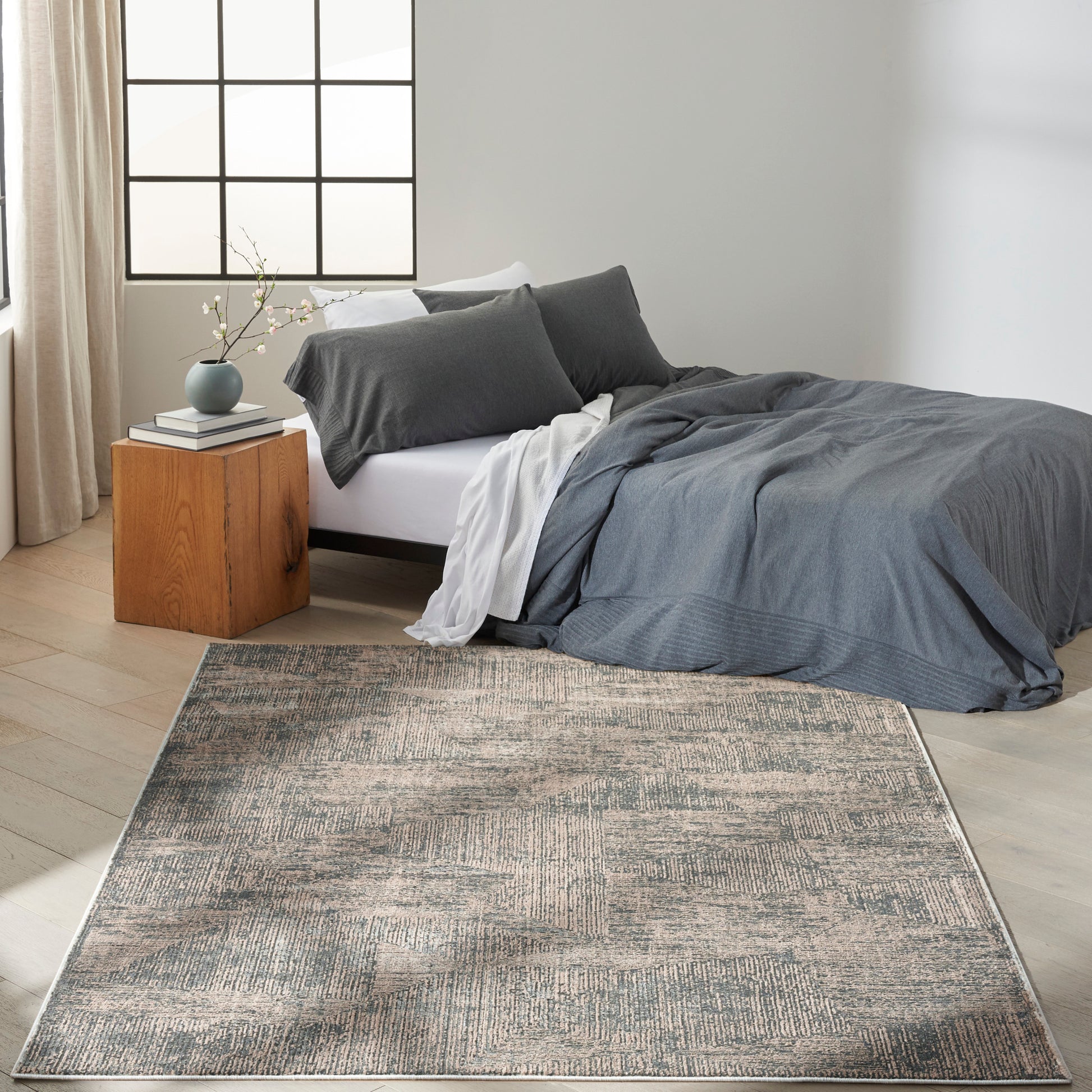 Cozy modern bedroom with dark gray duvet, gray pillows, white sheets, wooden cube nightstand, blue vase, beige curtains, rug.