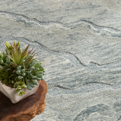 Succulent arrangement in a square concrete pot on a round wooden coaster, set on a blue-gray patterned rug.