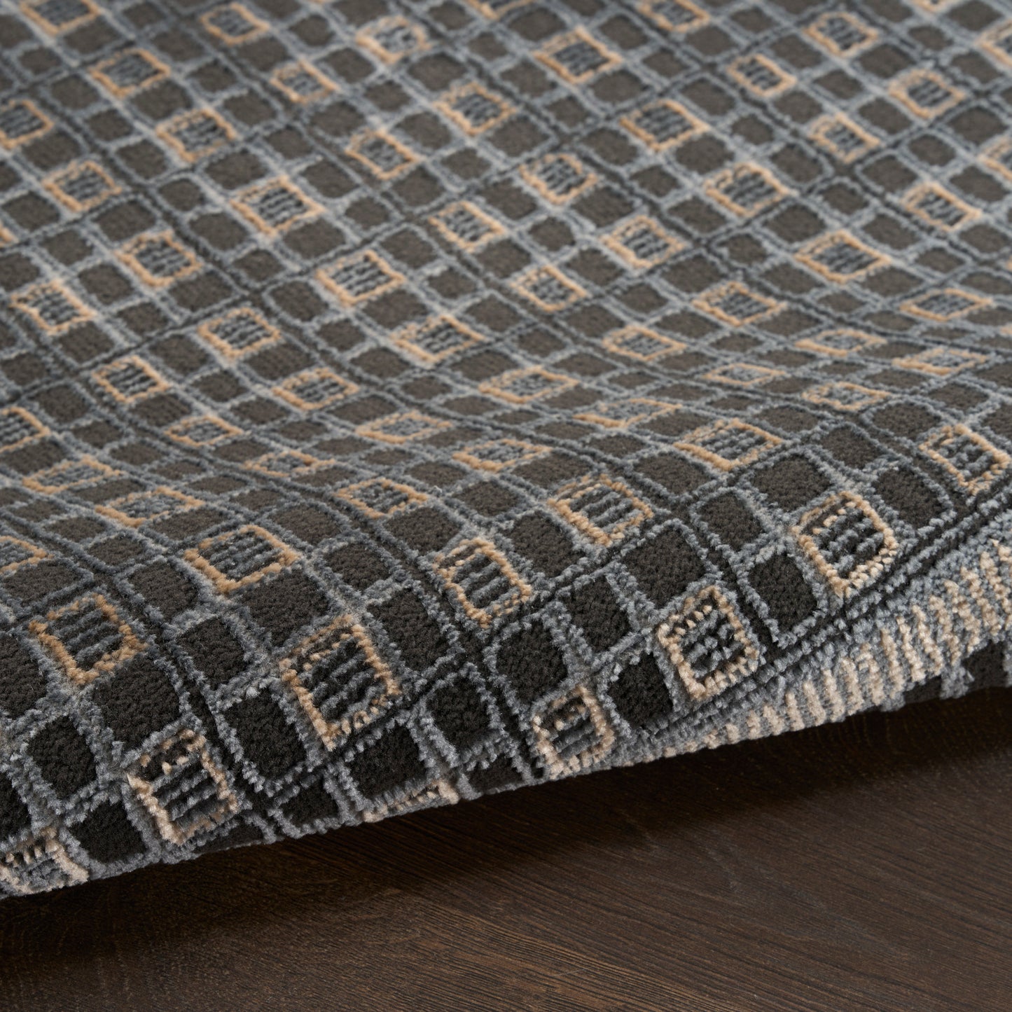 Dark gray woven rug with light gray and tan square-diamond geometric pattern, edge folded on a dark wood floor.
