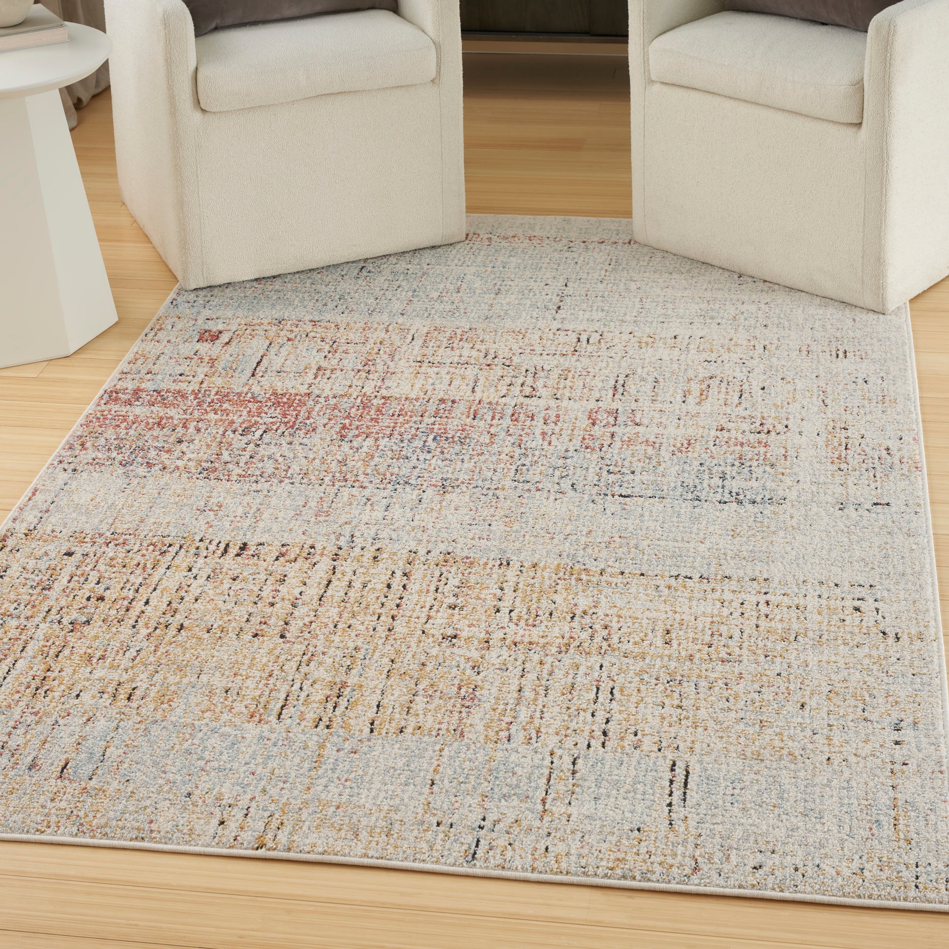 Rectangular woven area rug with abstract, distressed pattern in beige, tan, blue and pink; between cream armchairs.