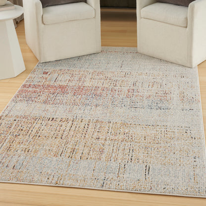 Rectangular woven area rug with abstract, distressed pattern in beige, tan, blue and pink; between cream armchairs.