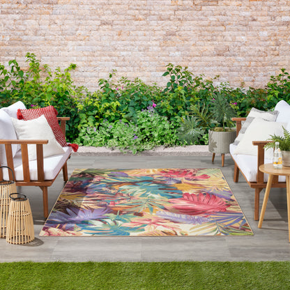 Outdoor patio set with white cushioned wooden chairs, vibrant tropical rug, greenery wall, lanterns & small side table.