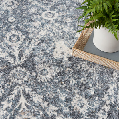 Blue-gray rug with an intricate white floral pattern; white planter with a green fern on a woven tray.