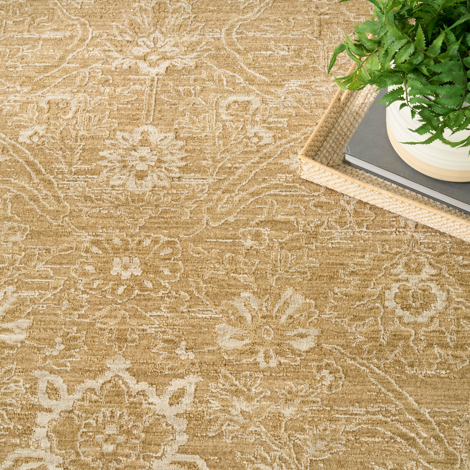 Beige floral-pattern rug with ornate motifs; on the top-right a wicker tray holding books and a potted green fern.