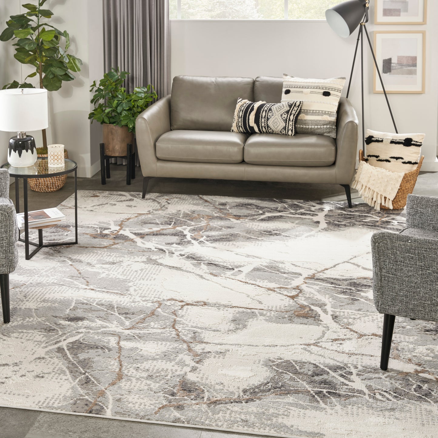 Taupe faux-leather sofa with black tapered legs in a modern living room, atop a gray abstract rug with plants, decorative cushions, and a tripod floor lamp.