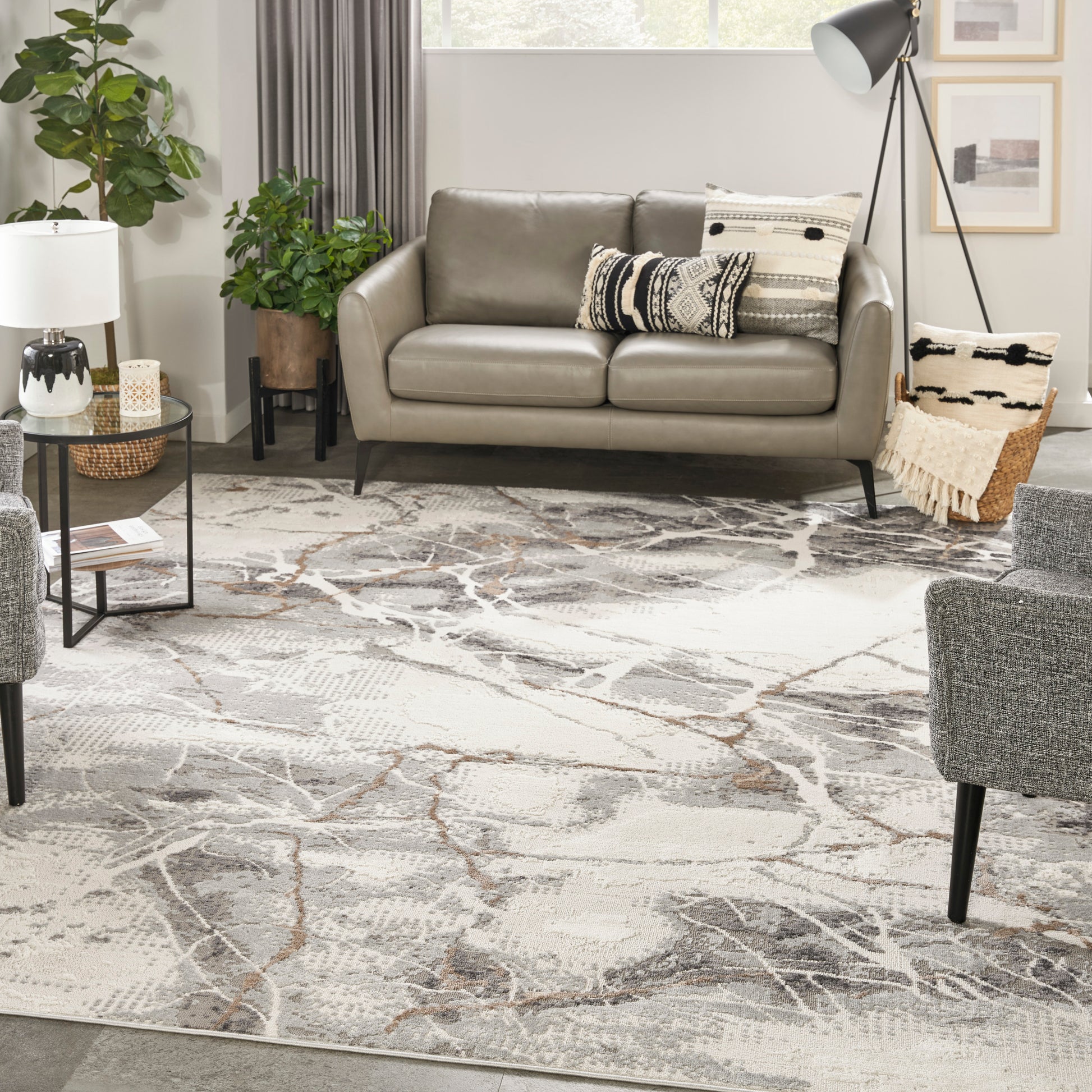 Taupe faux-leather sofa with black tapered legs in a modern living room, atop a gray abstract rug with plants, decorative cushions, and a tripod floor lamp.