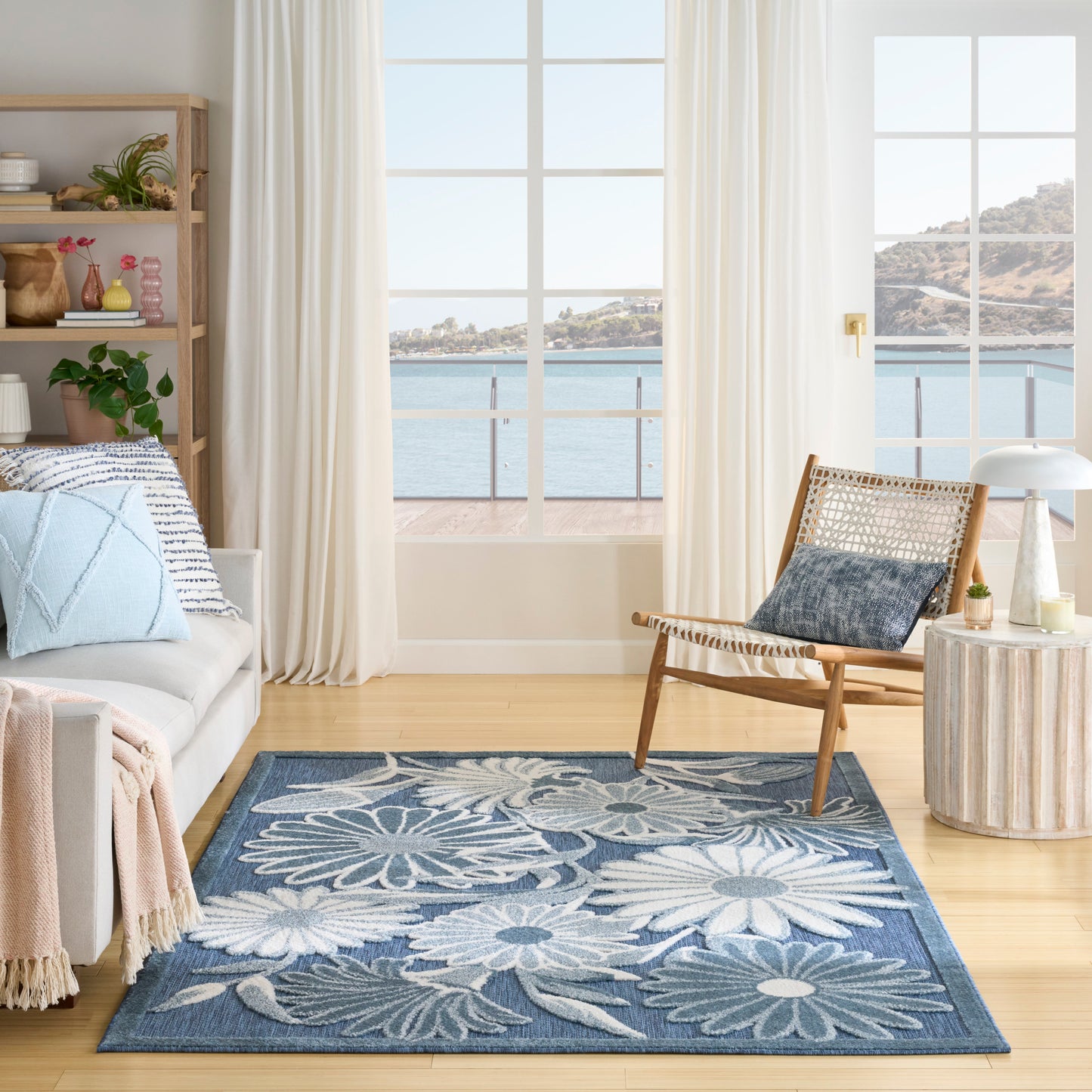 Coastal living room with blue floral rug, light wood floor, cream sofa, wicker chair, and ocean-view windows.