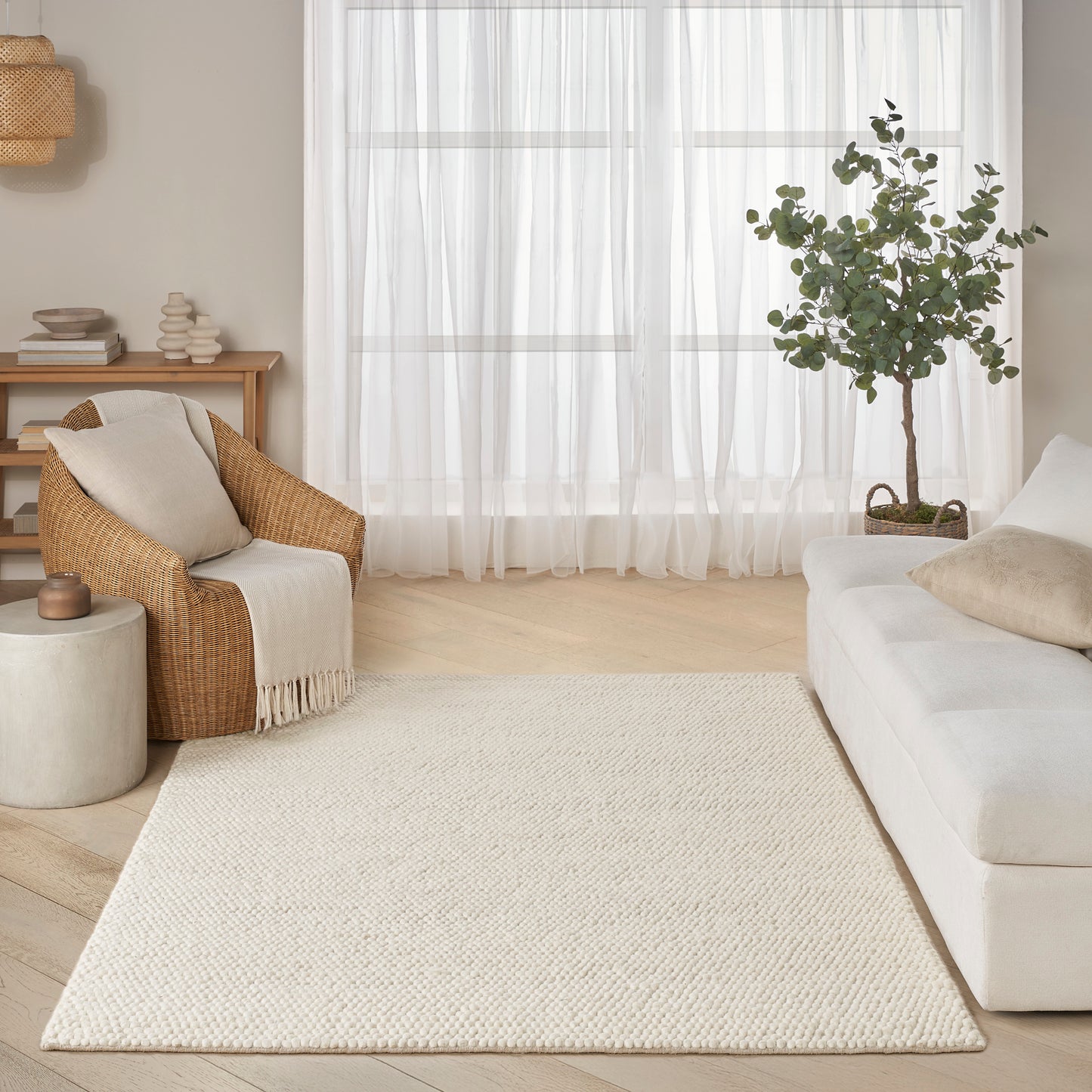 Bright living room with cream woven rug, white sofa, wicker armchair, potted tree, sheer curtains, and light wood accents.
