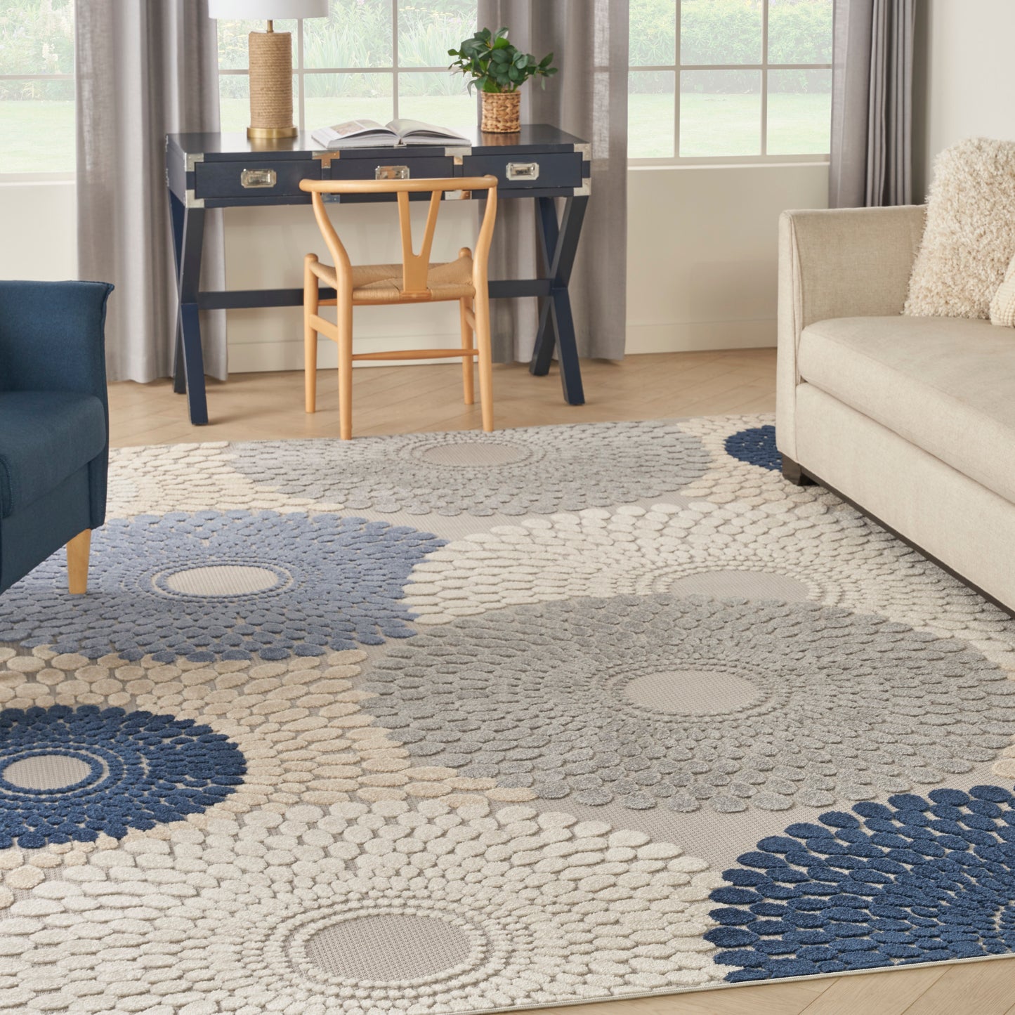 Living room with navy desk and wooden chair, large circular rosette rug in blue, gray and cream; blue armchair left, beige sofa right.