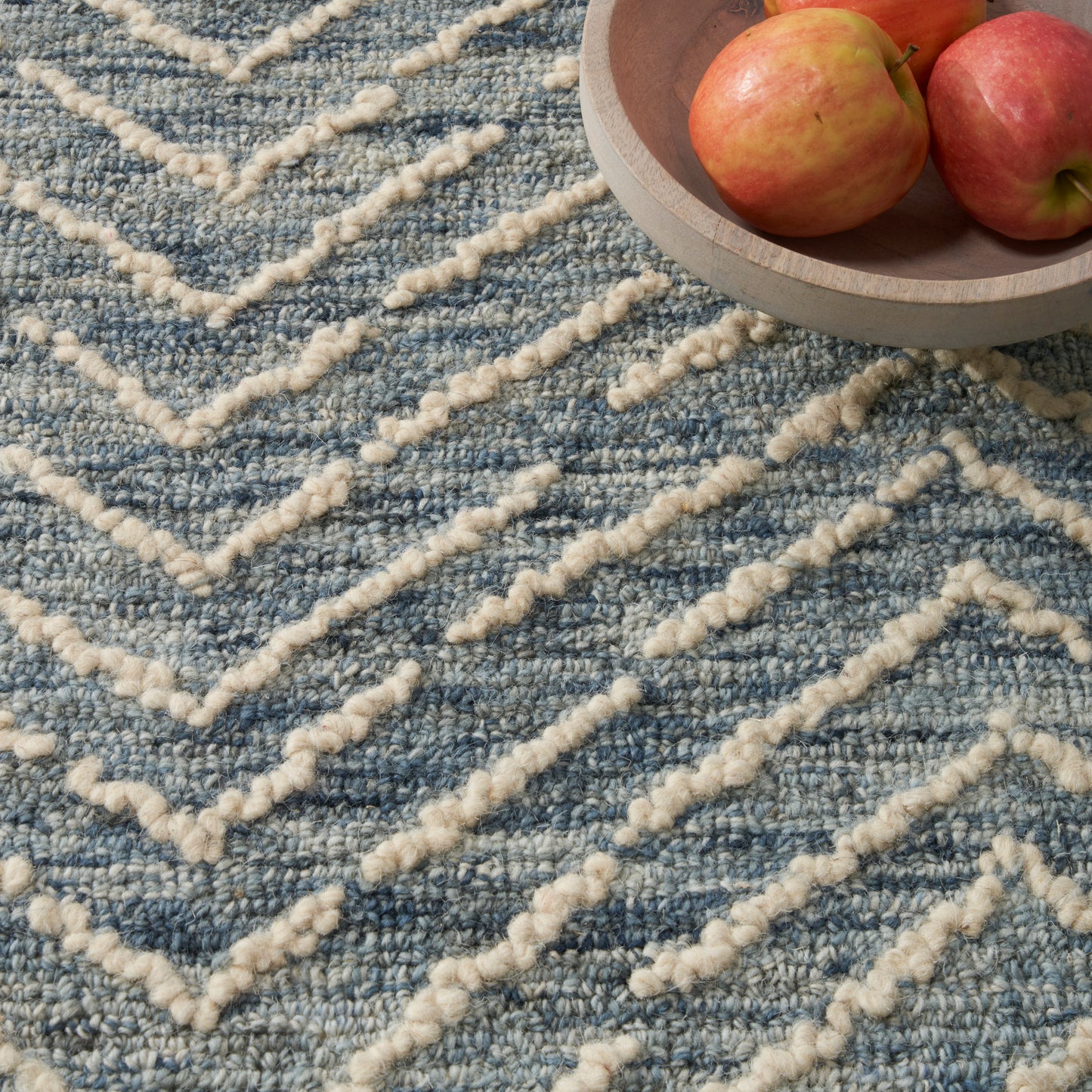 Blue textured rug with cream chunky chevron lines; round wooden bowl of apples resting in the corner.