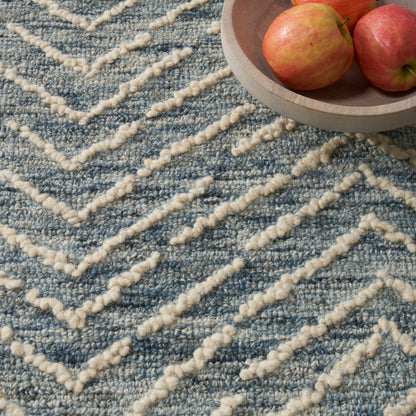 Blue textured rug with cream chunky chevron lines; round wooden bowl of apples resting in the corner.