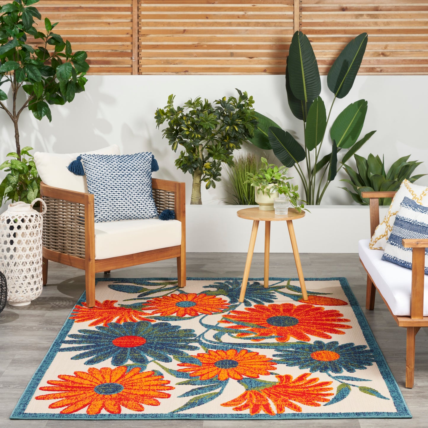Outdoor seating with wooden-frame white cushions, blue pillows, round wooden table, and a bright orange-blue floral rug.
