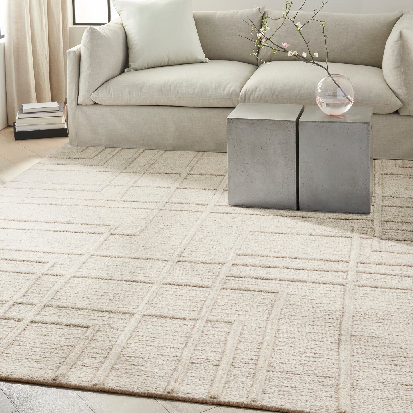 Beige geometric area rug with square-grid pattern in a modern living room; gray sofa, concrete block side tables, glass vase.