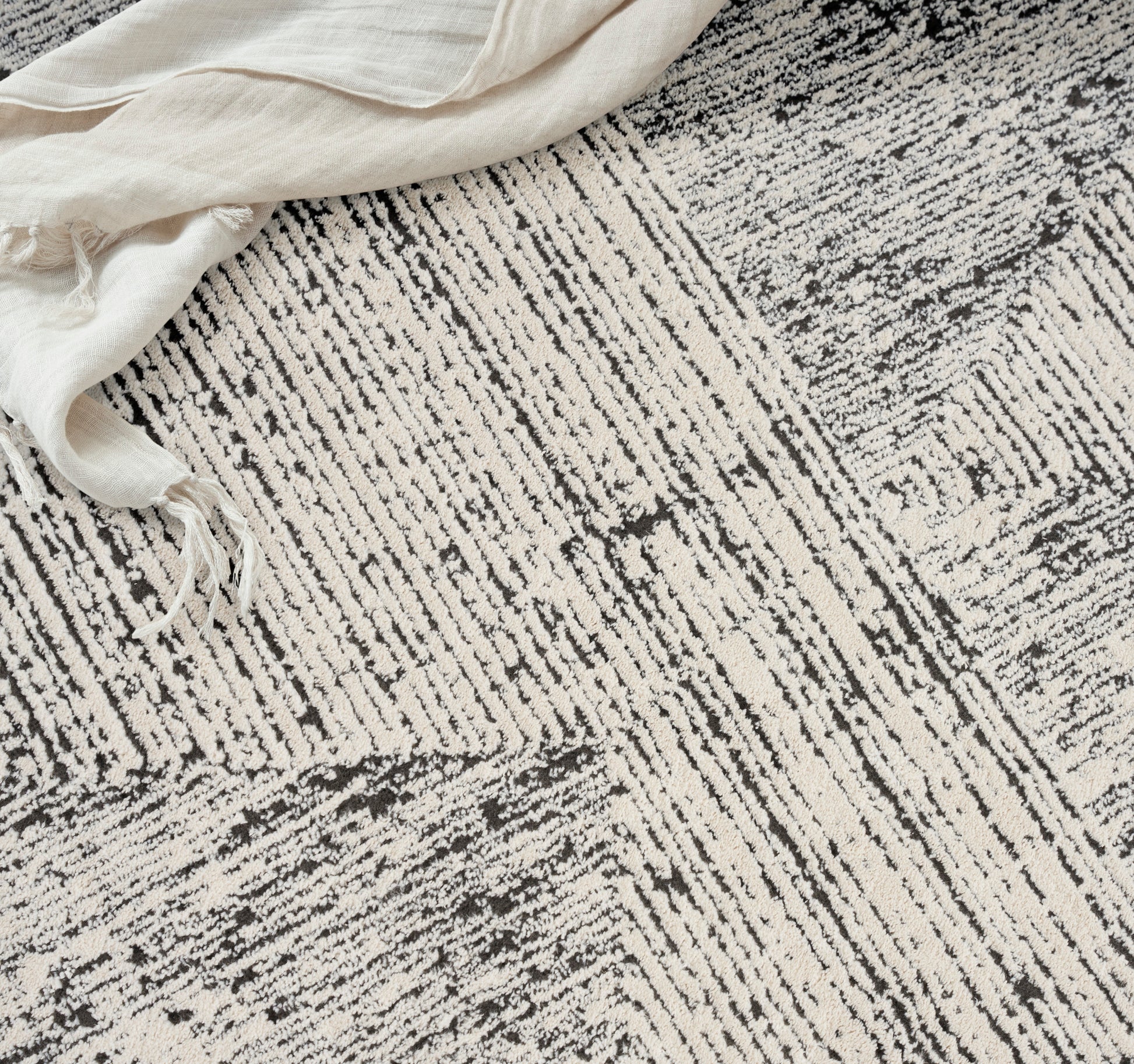 Beige-gray textured rug with irregular linear stripes and a cream linen cloth draped at the corner.