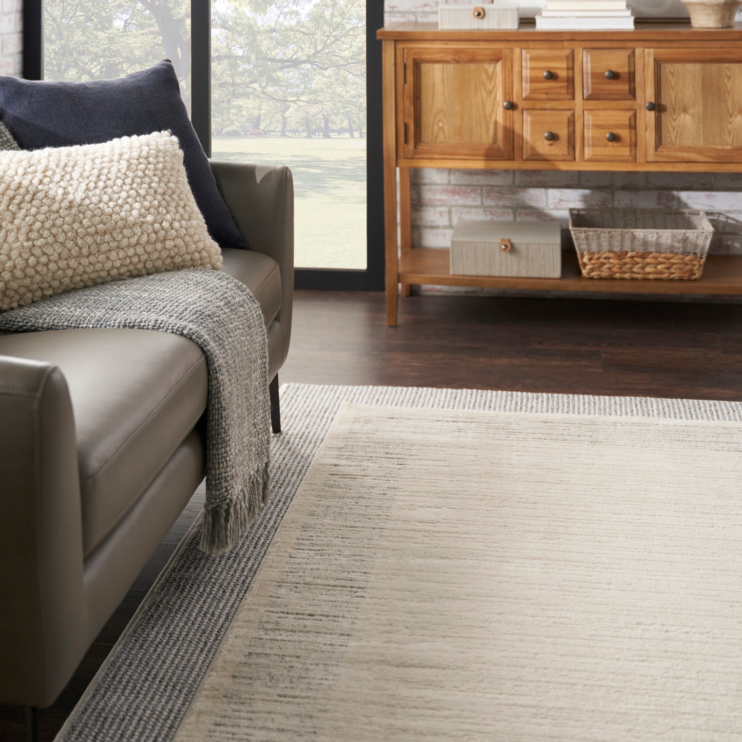 Living room with beige leather sofa, cream textured pillow, gray throw, light beige rug, and wooden sideboard with a basket.