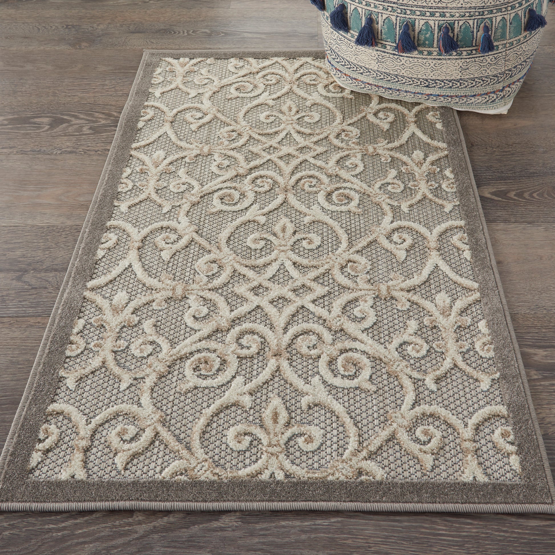 Beige area rug with ivory scrolling damask pattern, taupe background, bordered edges, on wooden floor beside a tasseled pouf.