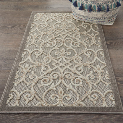 Beige area rug with ivory scrolling damask pattern, taupe background, bordered edges, on wooden floor beside a tasseled pouf.