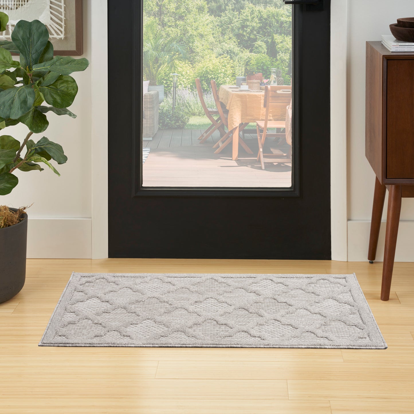 Light gray rectangular entryway rug with a subtle wavy pattern on wood floor; potted plant left, wooden cabinet right.