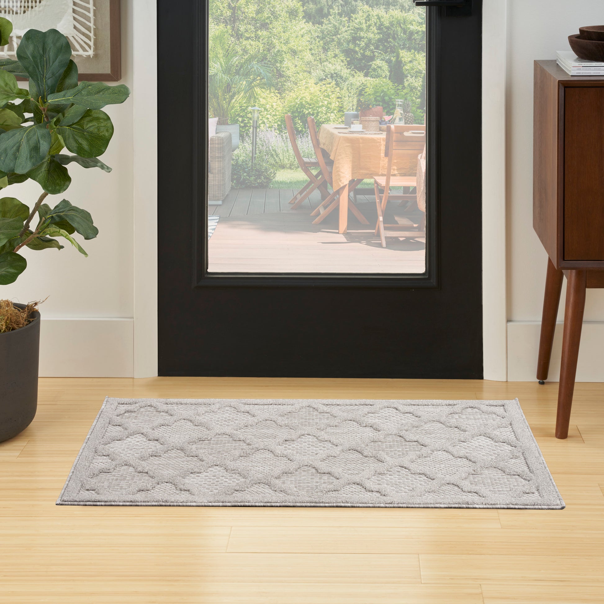 Light gray rectangular entryway rug with a subtle wavy pattern on wood floor; potted plant left, wooden cabinet right.