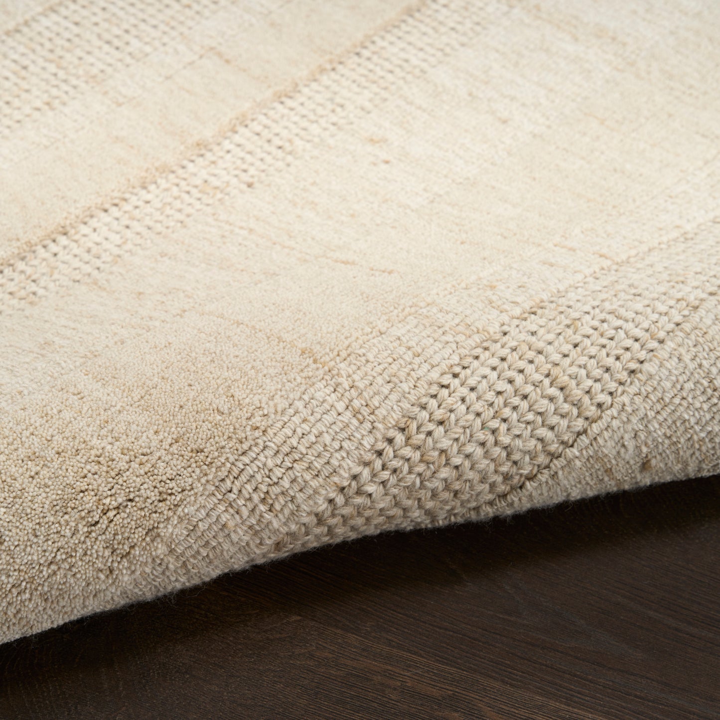 Beige textured rug close-up with woven patchwork pattern on dark wood floor.
