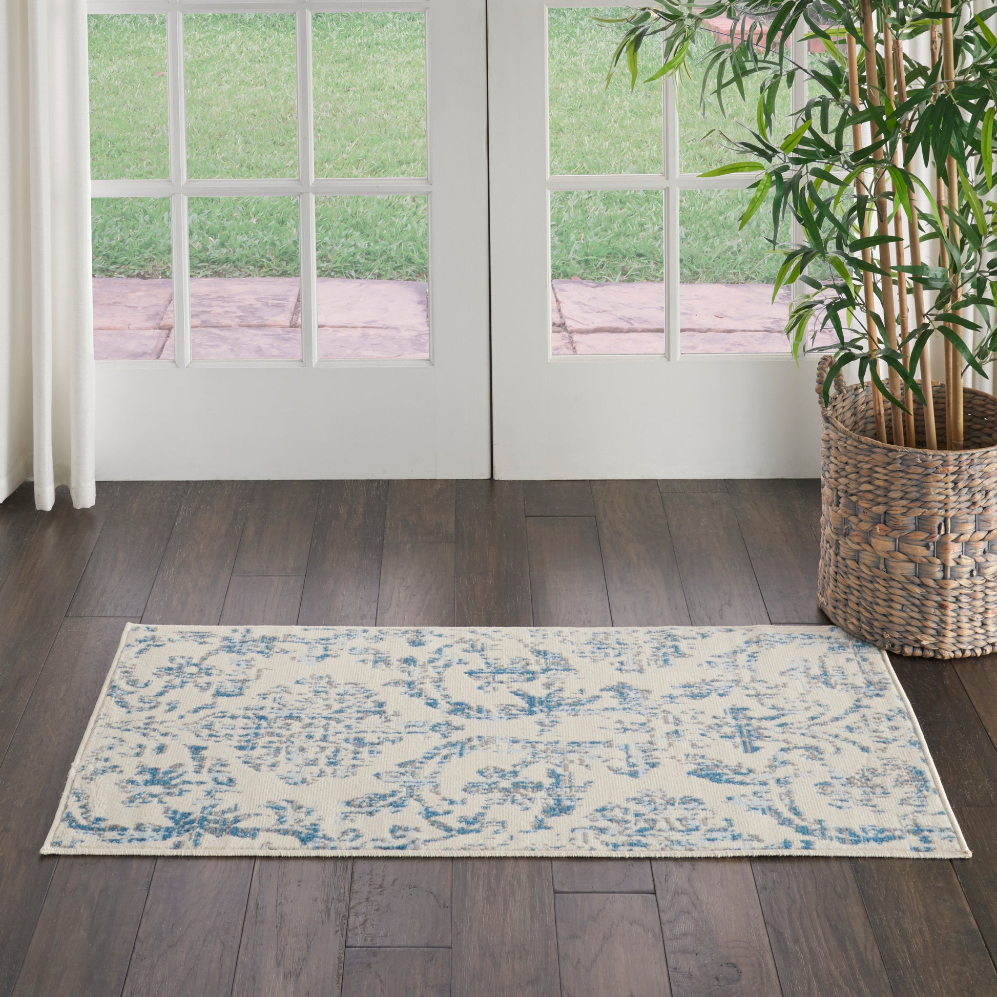 Light beige and blue patterned area rug on dark wood floor in front of French doors; potted plant in a woven basket to the right.