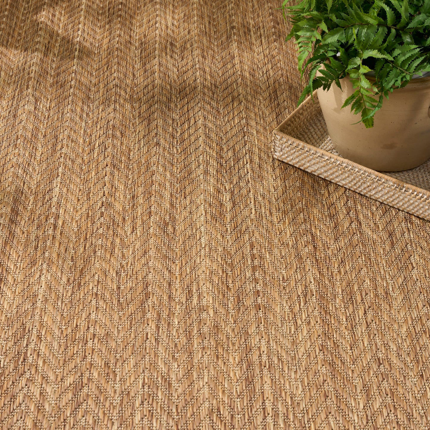 Natural jute rug with tan chevron pattern; green fern in clay pot on a woven tray.