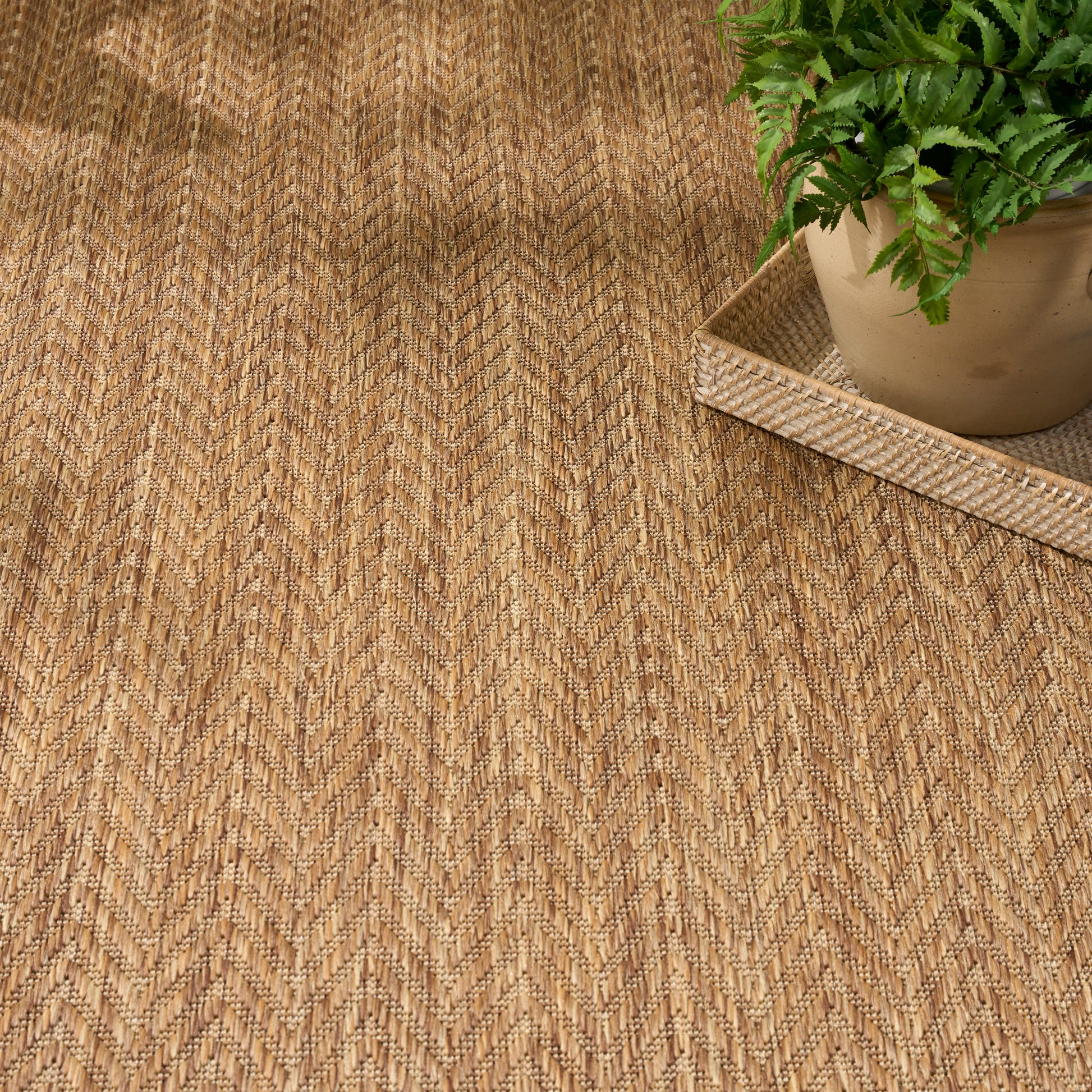 Natural jute rug with tan chevron pattern; green fern in clay pot on a woven tray.