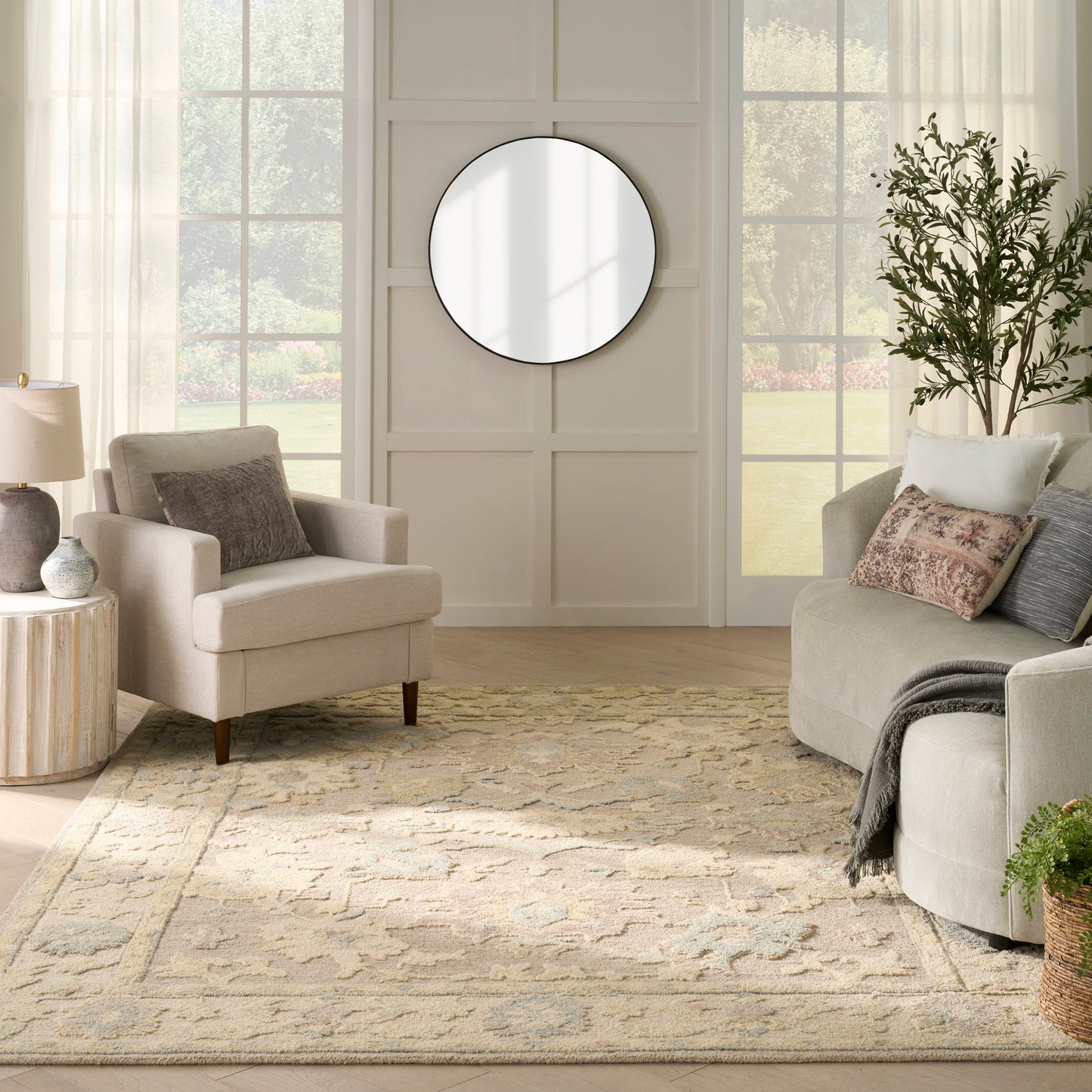 Neutral living room with beige sofa and armchair, round wall mirror, cream patterned rug, tall plant.