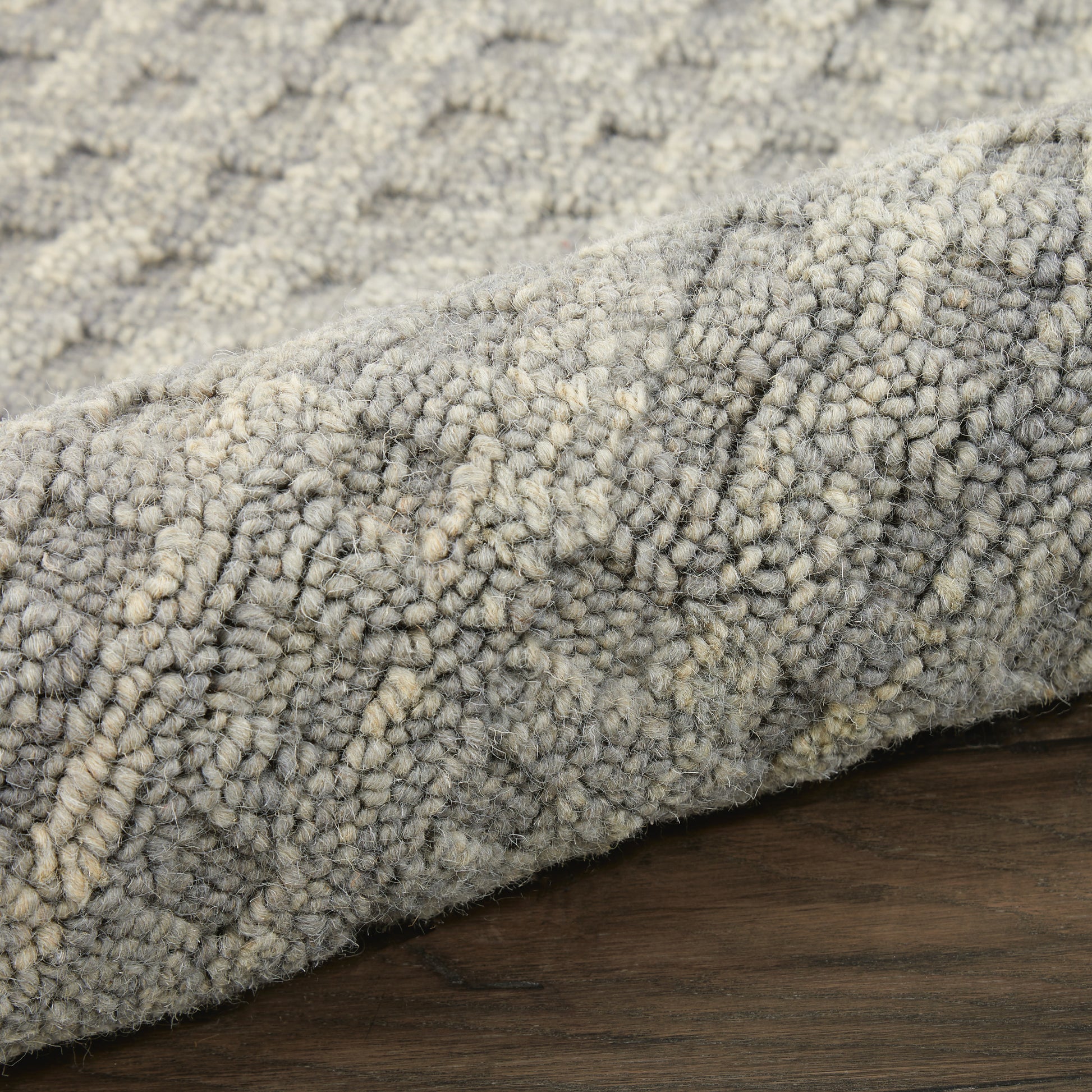 Close-up of a gray and ivory loop-pile rug with dense textured fibers, rolled edge on a dark wooden floor.