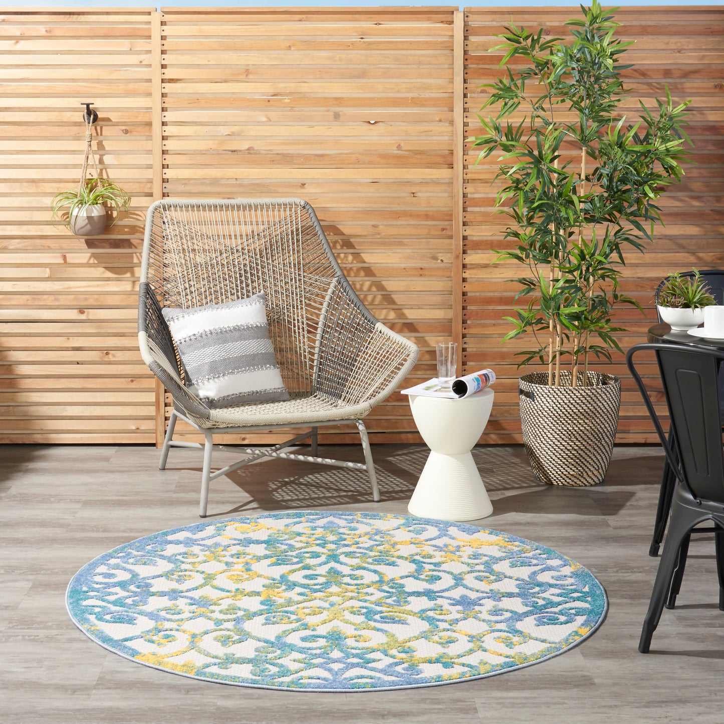 Outdoor patio with wicker chair, striped cushion, round blue-yellow patterned rug, potted plants and a wooden fence.