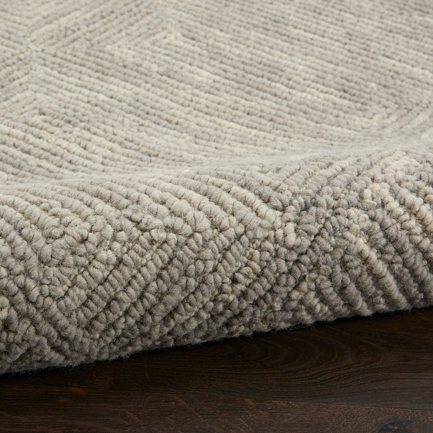 Beige thick-pile rug with geometric diamond pattern, rolled edge on dark wooden floor.