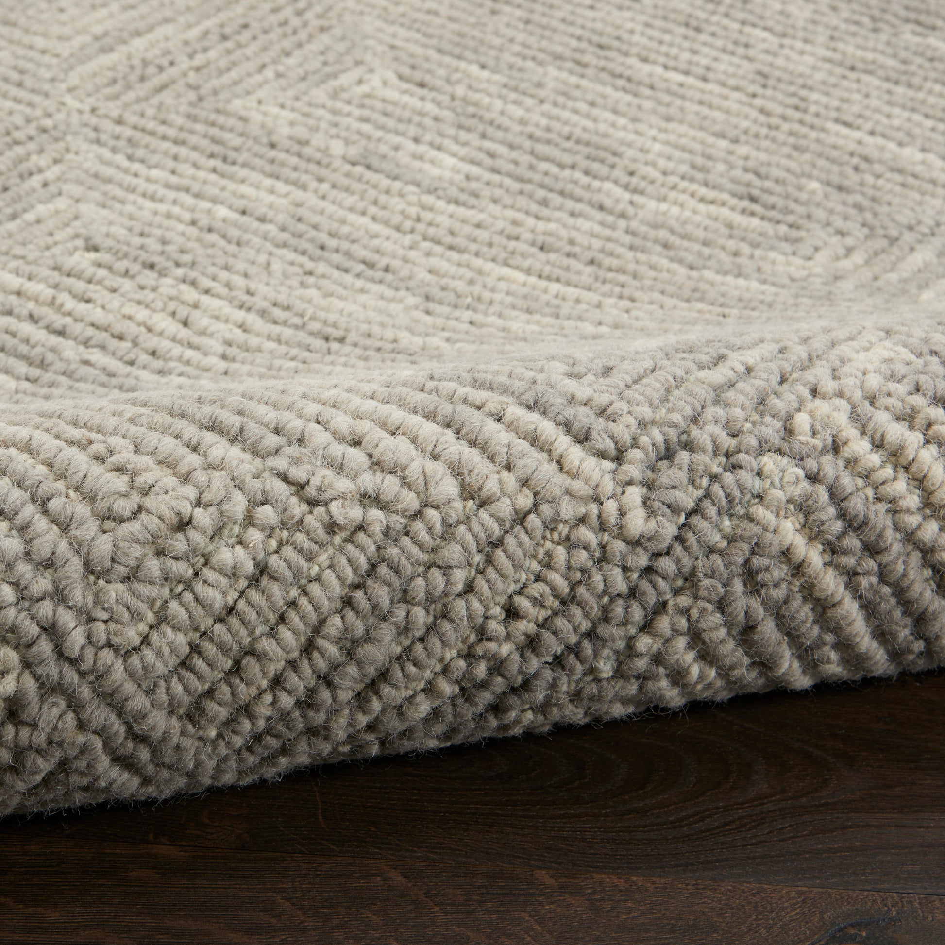 Beige thick-pile rug with geometric diamond pattern, rolled edge on dark wooden floor.
