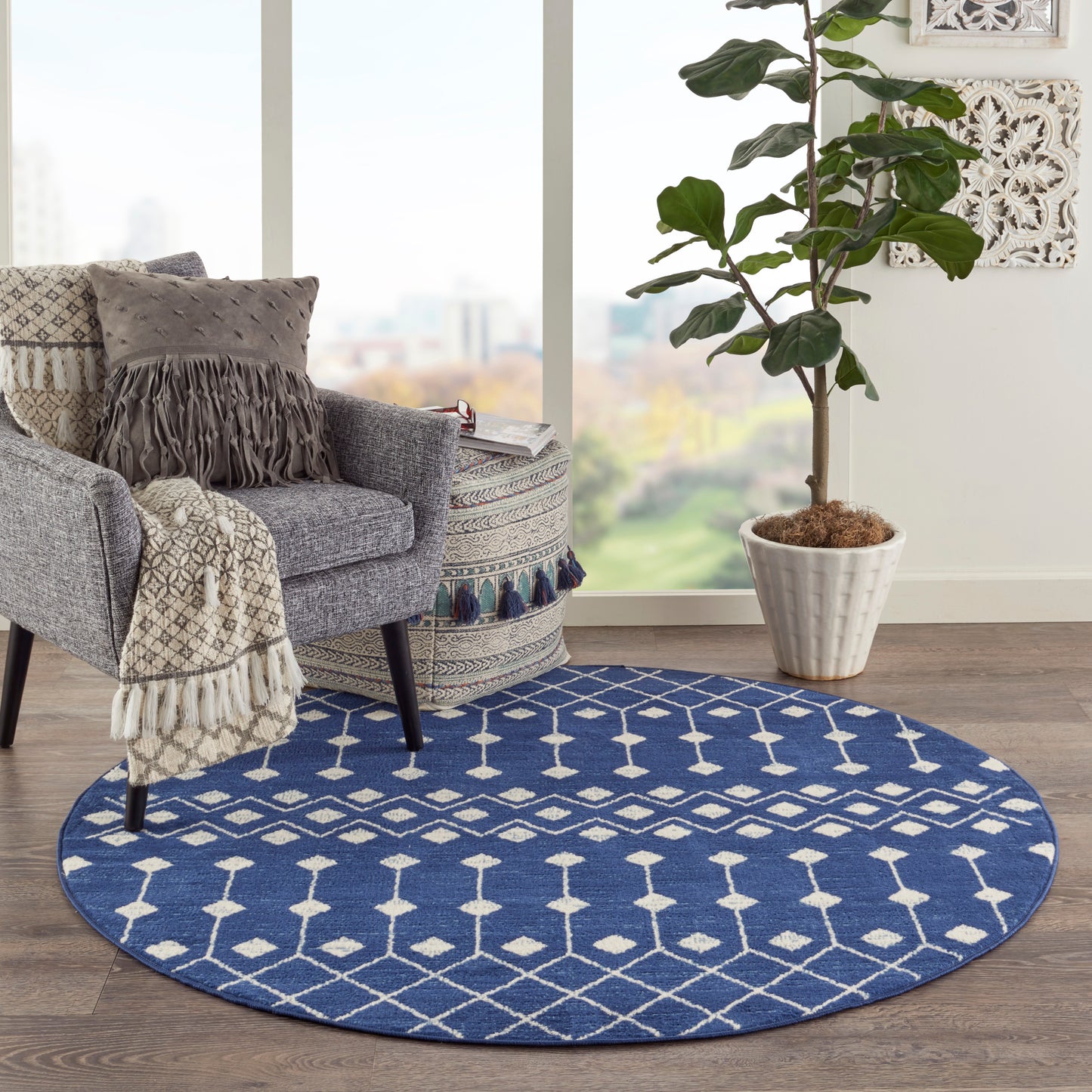 Blue round rug with white geometric diamonds under a gray armchair, fringe throw, potted plant by a sunny window.