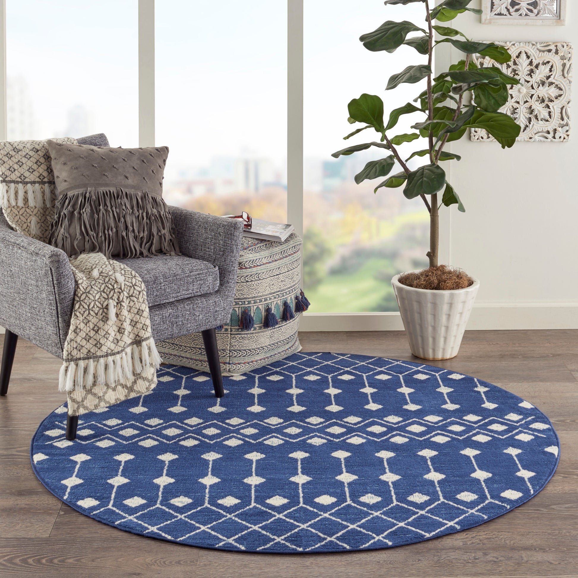 Blue round rug with white geometric diamonds under a gray armchair, fringe throw, potted plant by a sunny window.