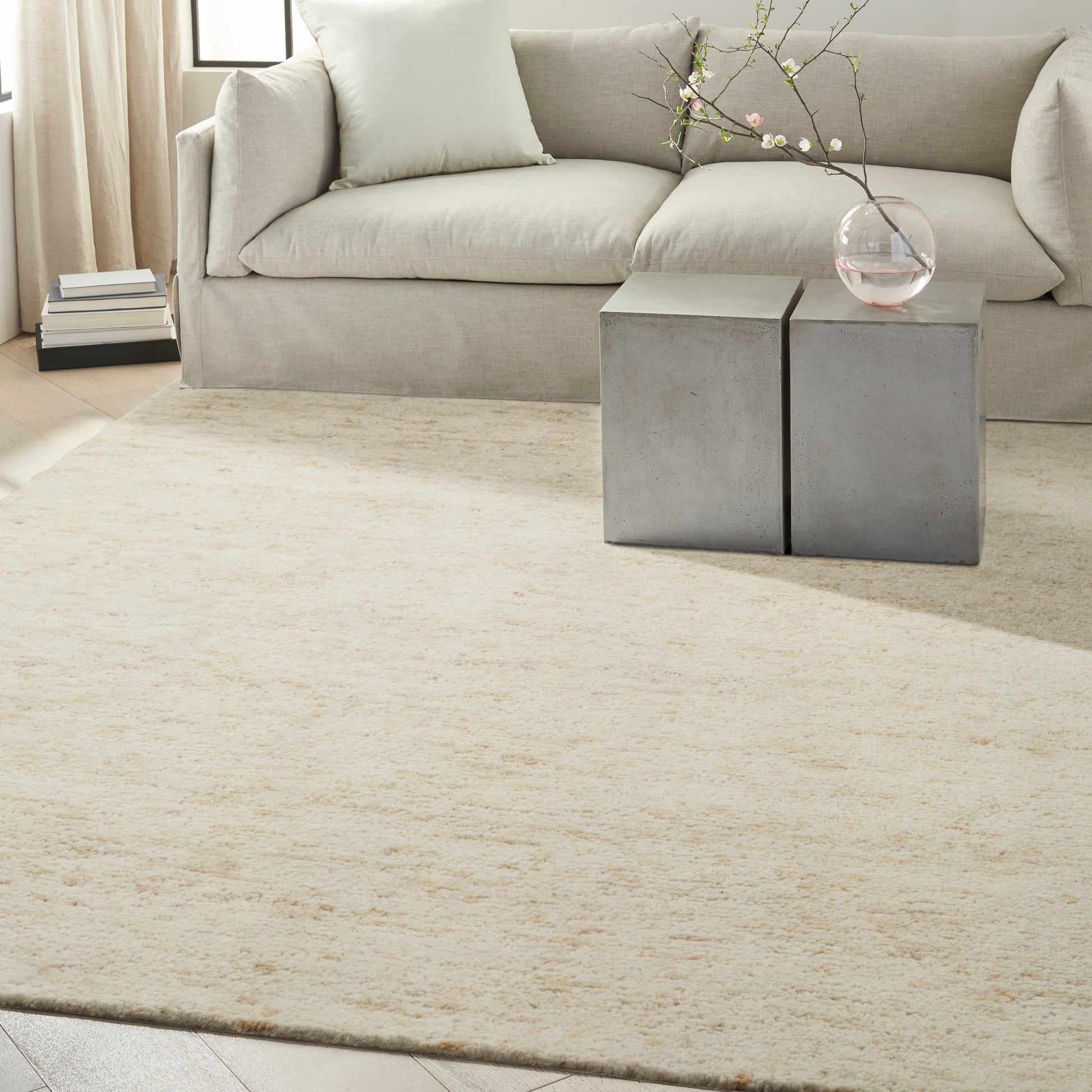 Neutral living room: cream-beige rug, light gray sofa, pair of square concrete side tables, glass vase with branch.