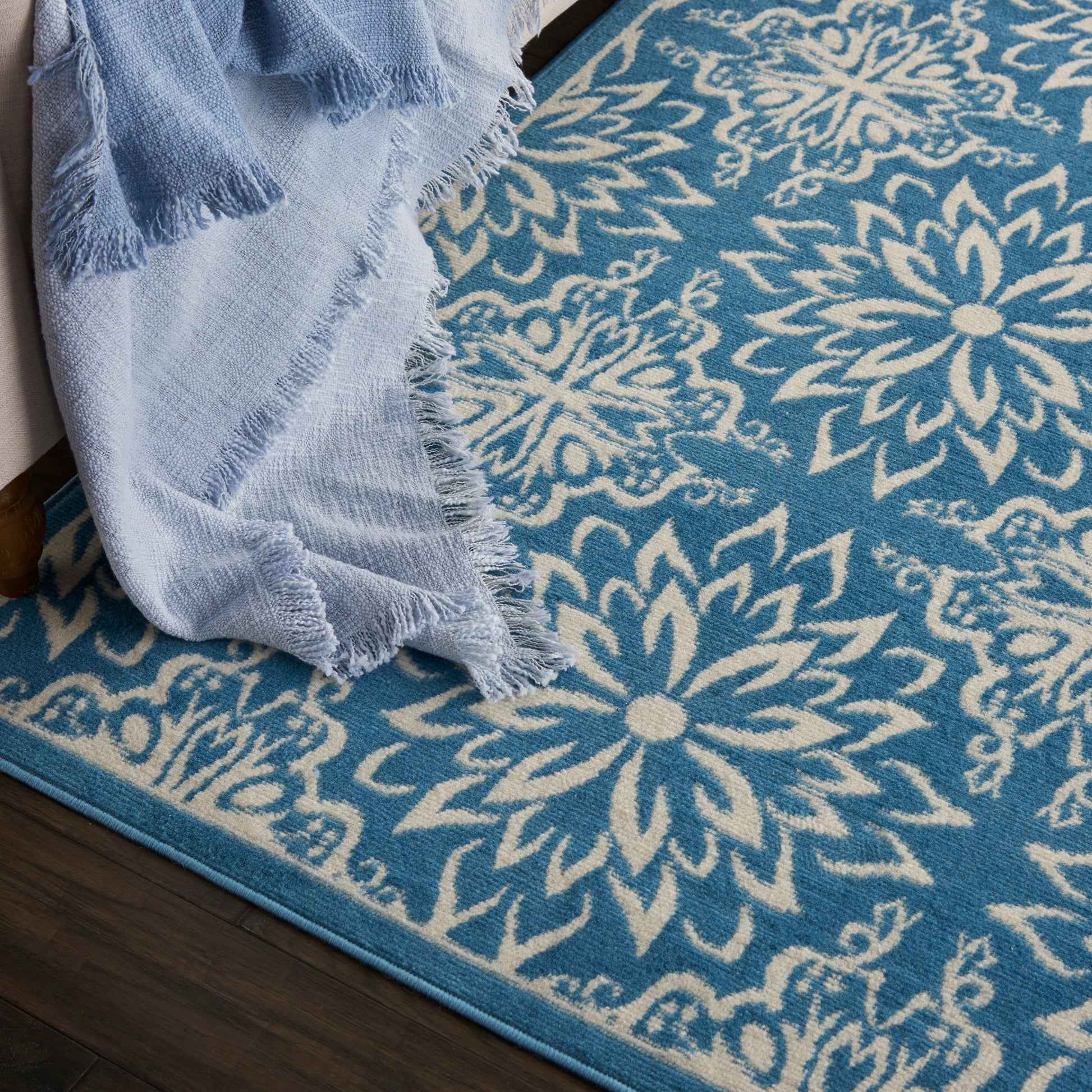 Blue area rug with large white floral medallion pattern, turquoise border; light blue fringed blanket draped along edge.