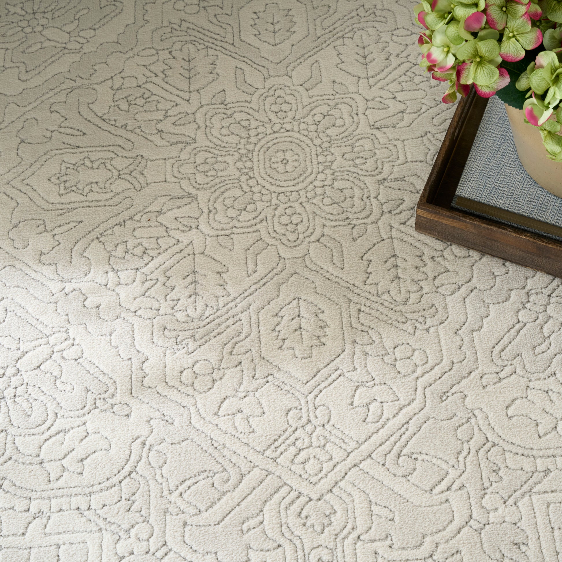 Cream damask patterned rug with raised floral motif; corner plant in a wooden tray adds natural contrast.