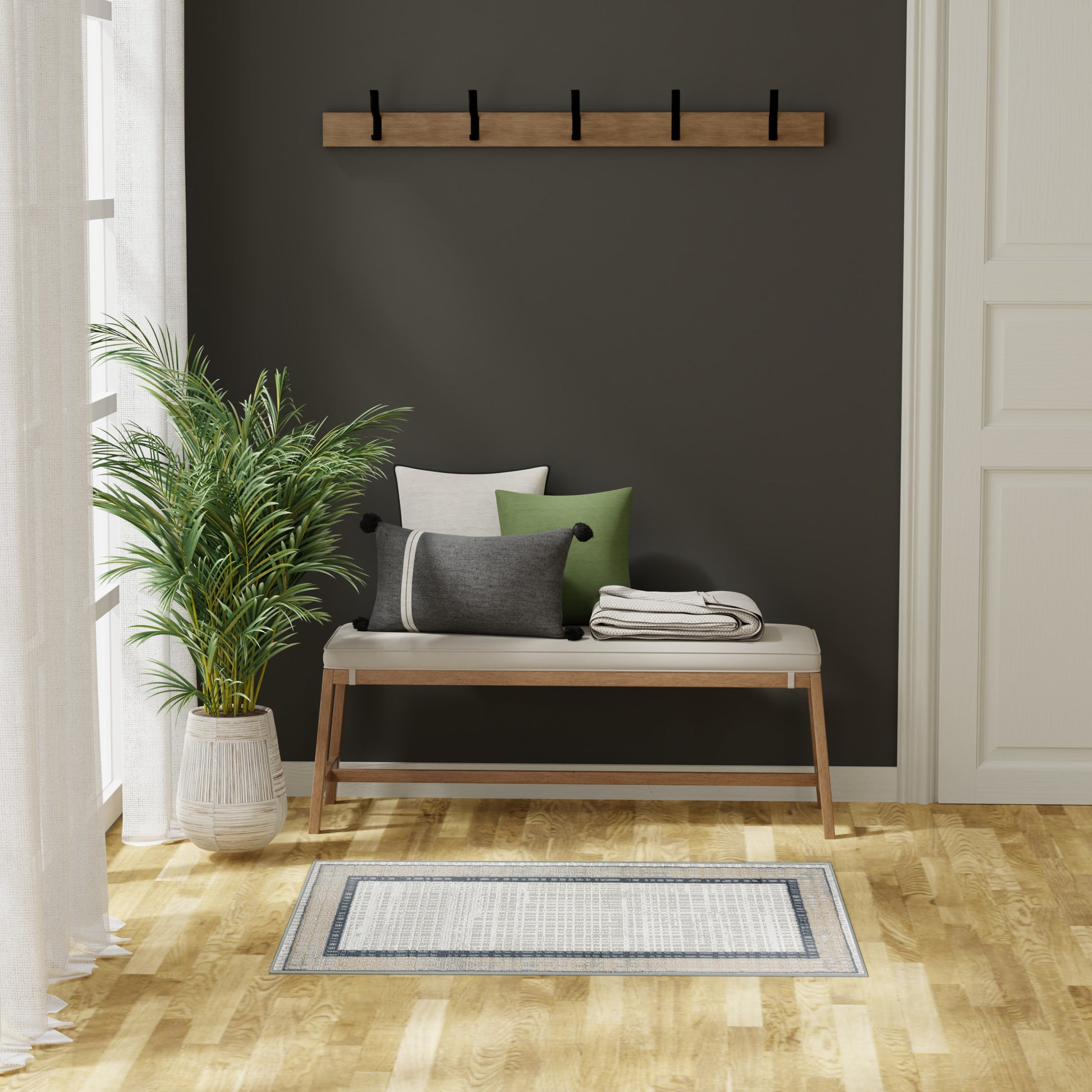 Wood bench with beige cushion & gray/green pillows, black hook rack, potted palm, light hardwood floor, striped rug