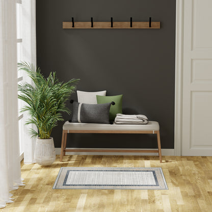 Wood bench with beige cushion & gray/green pillows, black hook rack, potted palm, light hardwood floor, striped rug