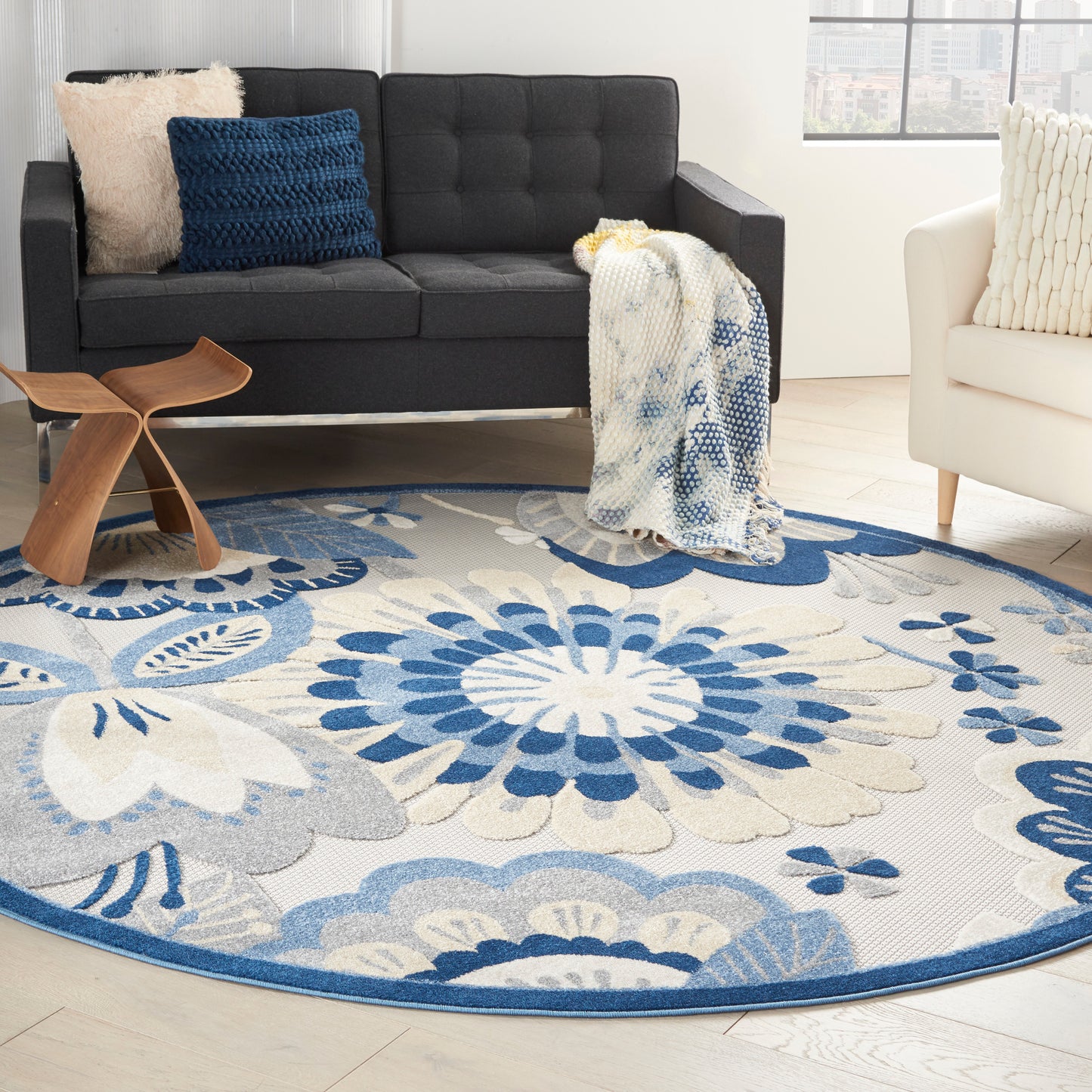 Round blue-and-white floral area rug in a modern living room with a dark gray sofa, white chair, and cozy throw blanket.
