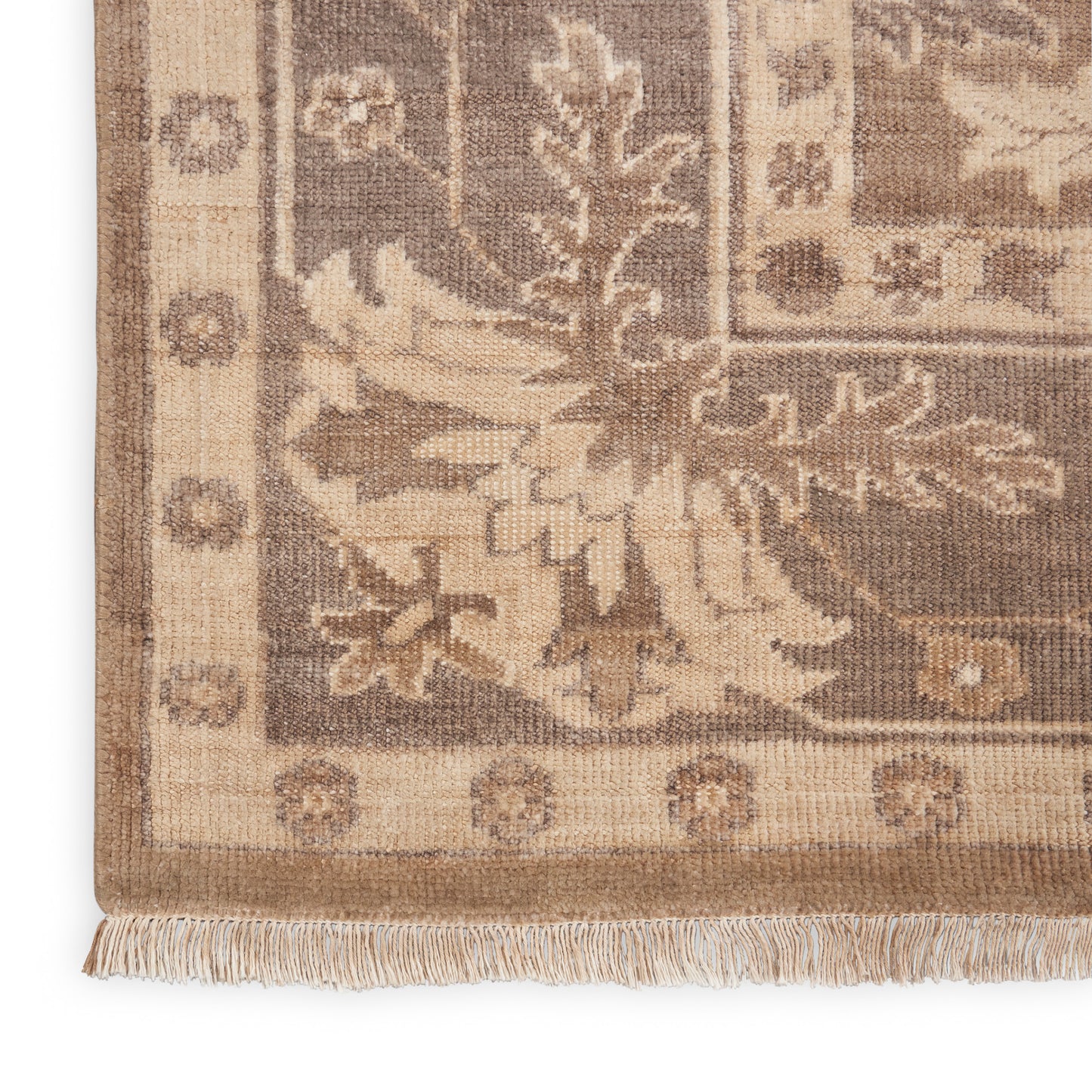 Beige-brown woven rug corner with floral pattern, ornate border and fringe along the bottom edge.