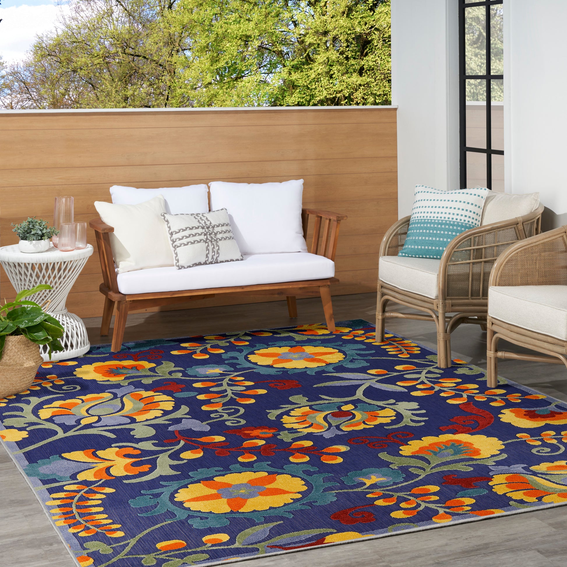 Outdoor patio with navy floral rug and yellow/orange flowers; white-cushion wooden loveseat, wicker chairs, and side table.