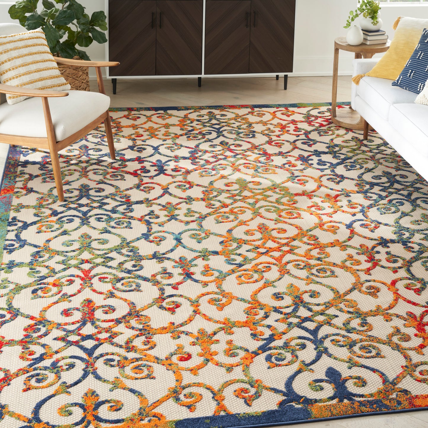 Living room with a vibrant multicolor floral scroll rug in blues, oranges & teals; mid-century chairs, white sofa with yellow/blue cushions, wood cabinet.
