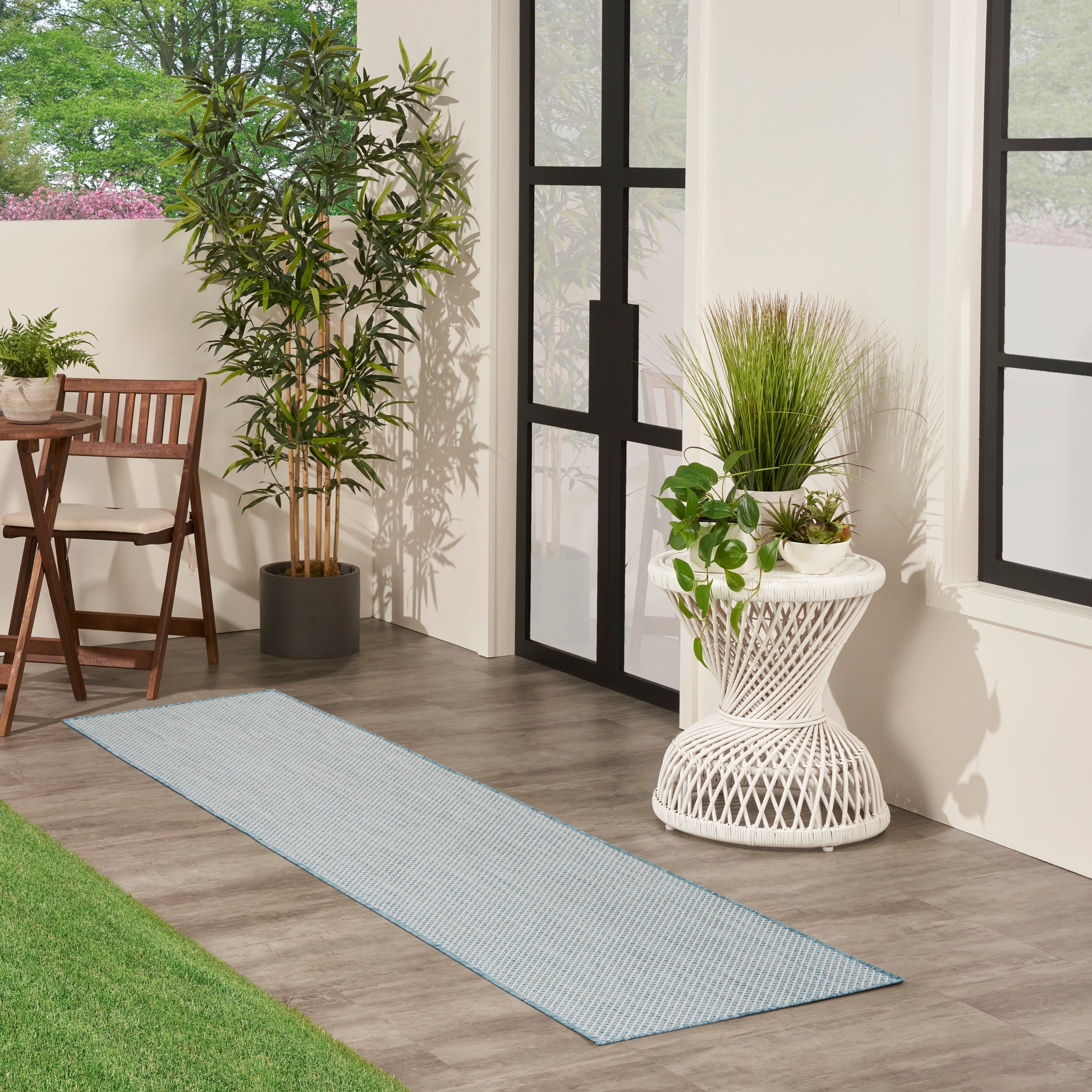 Long light-blue woven runner rug on wood floor beside potted plants and a white wicker plant stand.