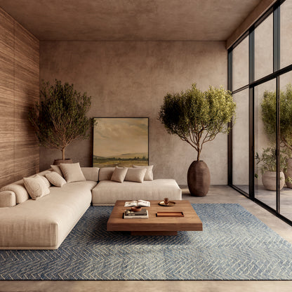 Beige sectional sofa with neutral cushions, blue zigzag rug, wooden coffee table, large potted trees, earthy walls, and glass wall windows.