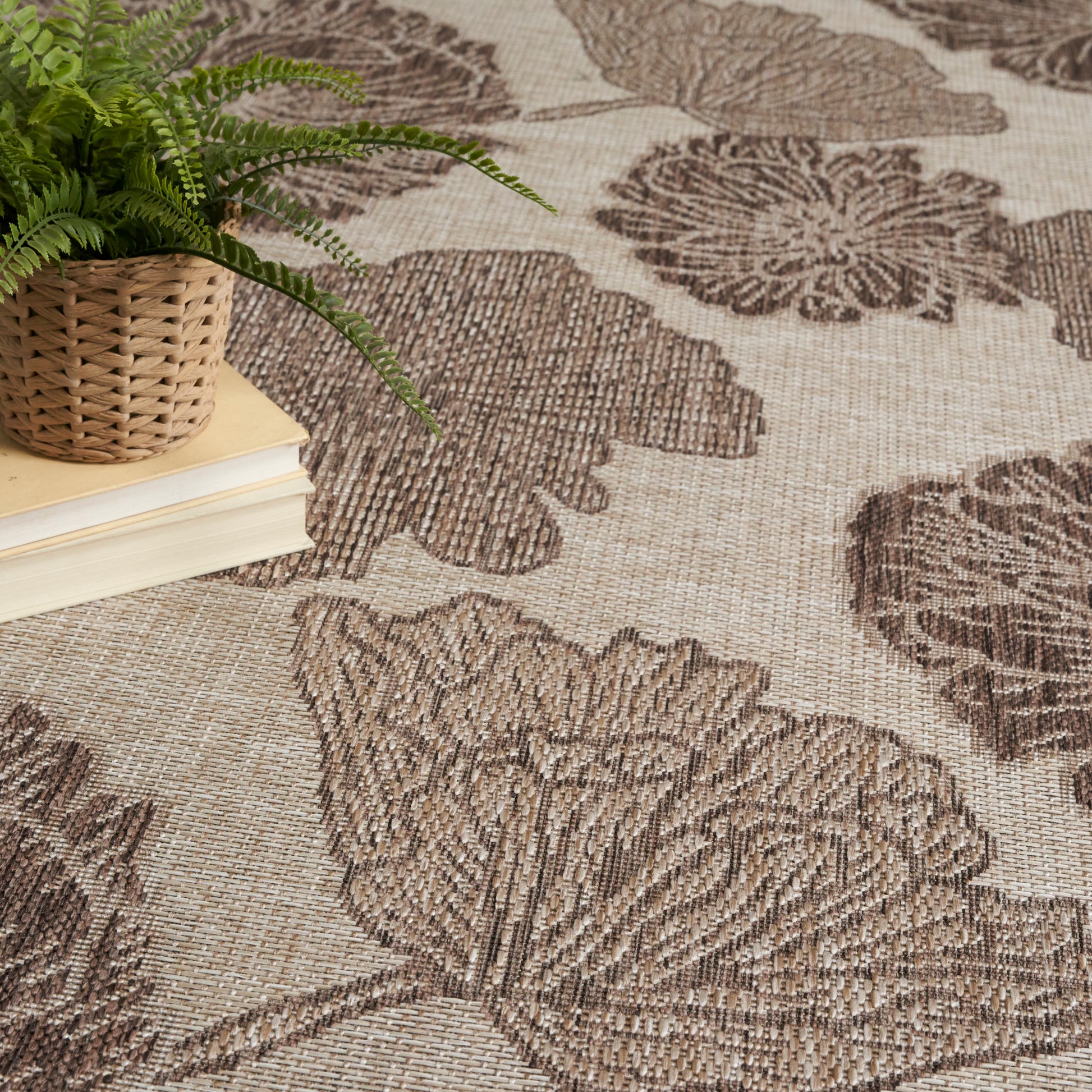 Beige woven rug with a large brown botanical leaf pattern; a potted fern in a wicker basket rests on a stack of books.