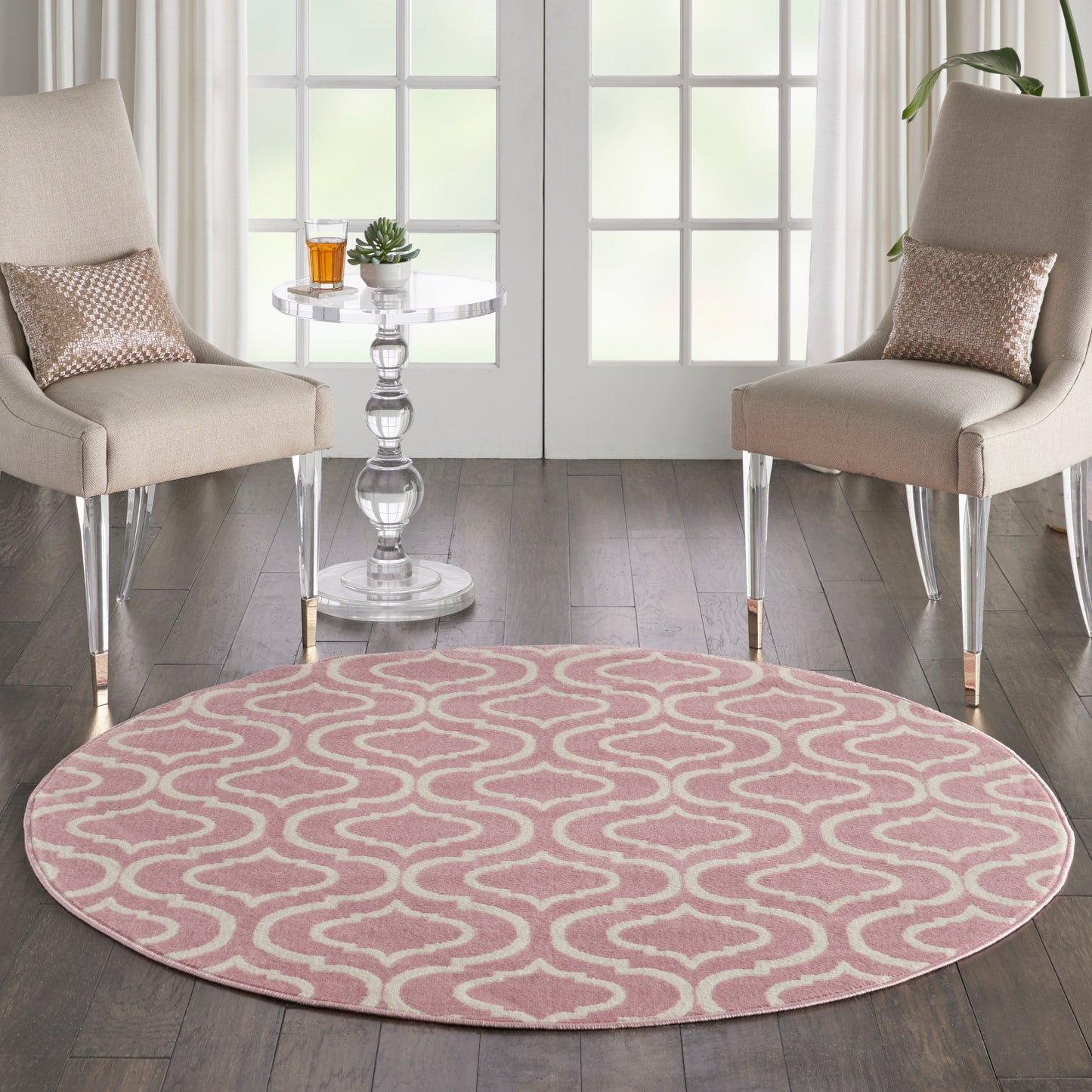 Round pink rug with white wave pattern between two beige upholstered chairs and a glass pedestal side table in a living room.
