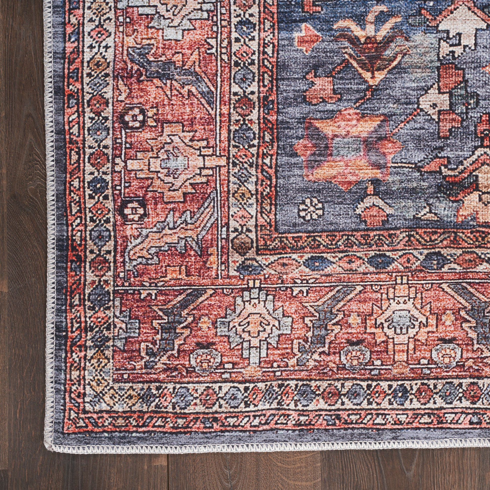 Woven wool oriental rug corner with red and navy patterns, ivory accents, fringe along edge on dark wood floor.