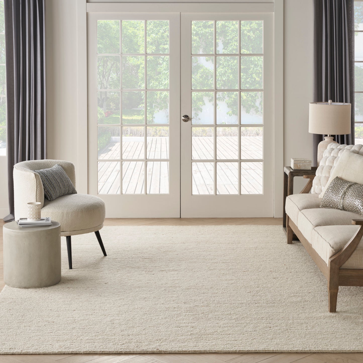 Neutral living room with white-framed French doors, beige sofa, curved cream chair, cream rug, side tables, gray curtains.