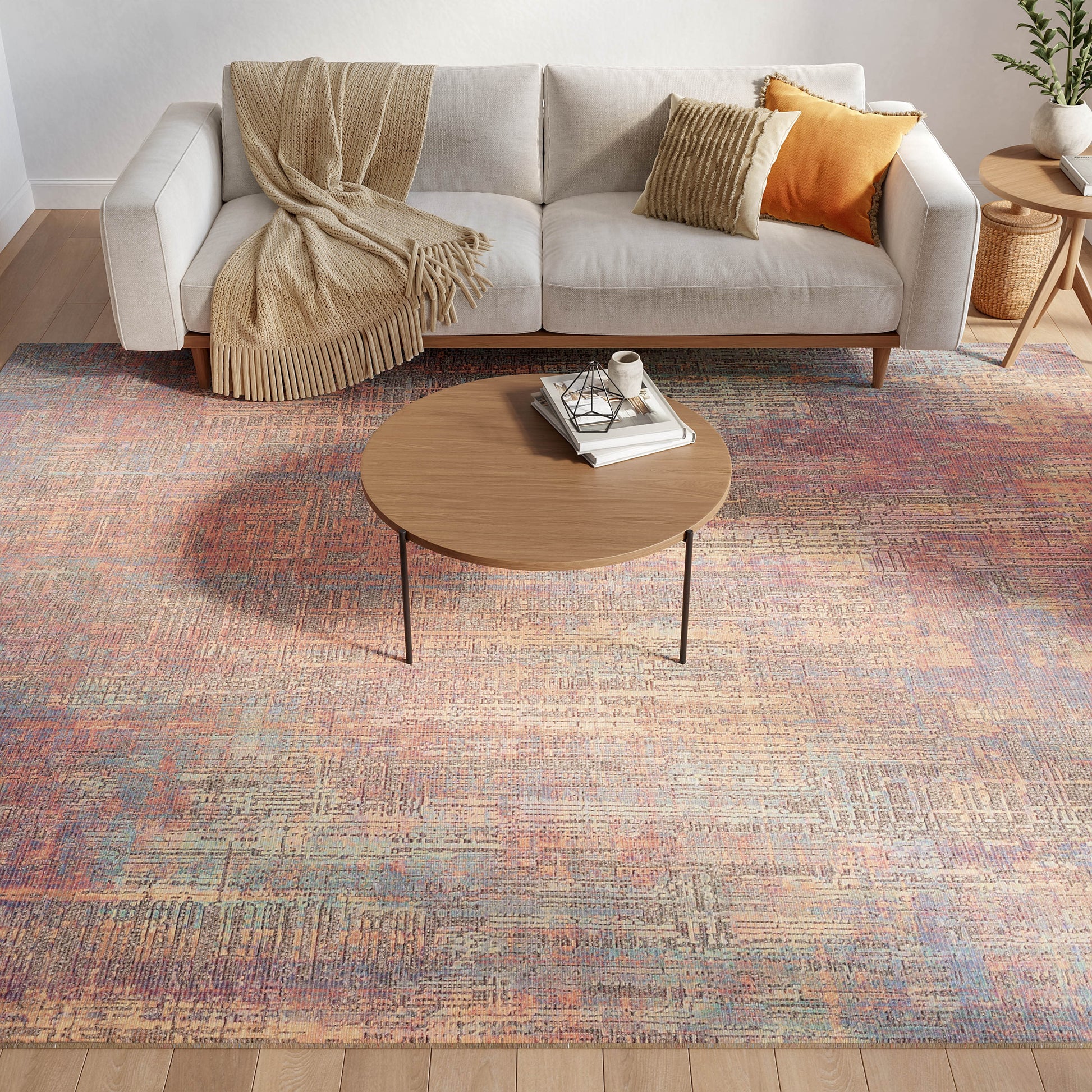 Light gray sofa with beige throw and orange and yellow cushions, wooden coffee table, colorful abstract rug, side plant.